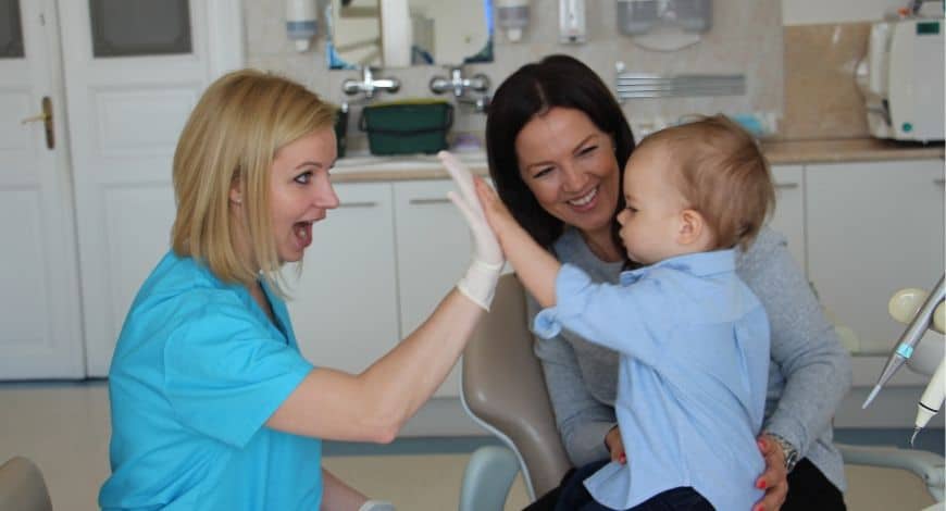 Dr. Friedreich Dóra elégedett, mosolygó páciens ölében tartott gyermekkel pacsizik az Artdent Fogászat rendelőjében